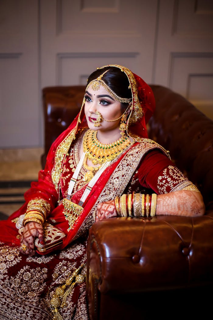 services-02 South Asian bride in traditional red and gold attire with henna and jewelry indoors.